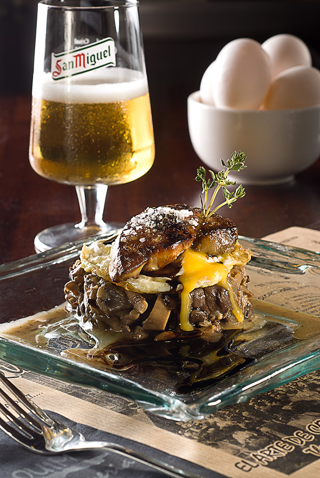 Hamburguesa con huevo, cerveza y un plato con huevos sobre una mesa rústica.